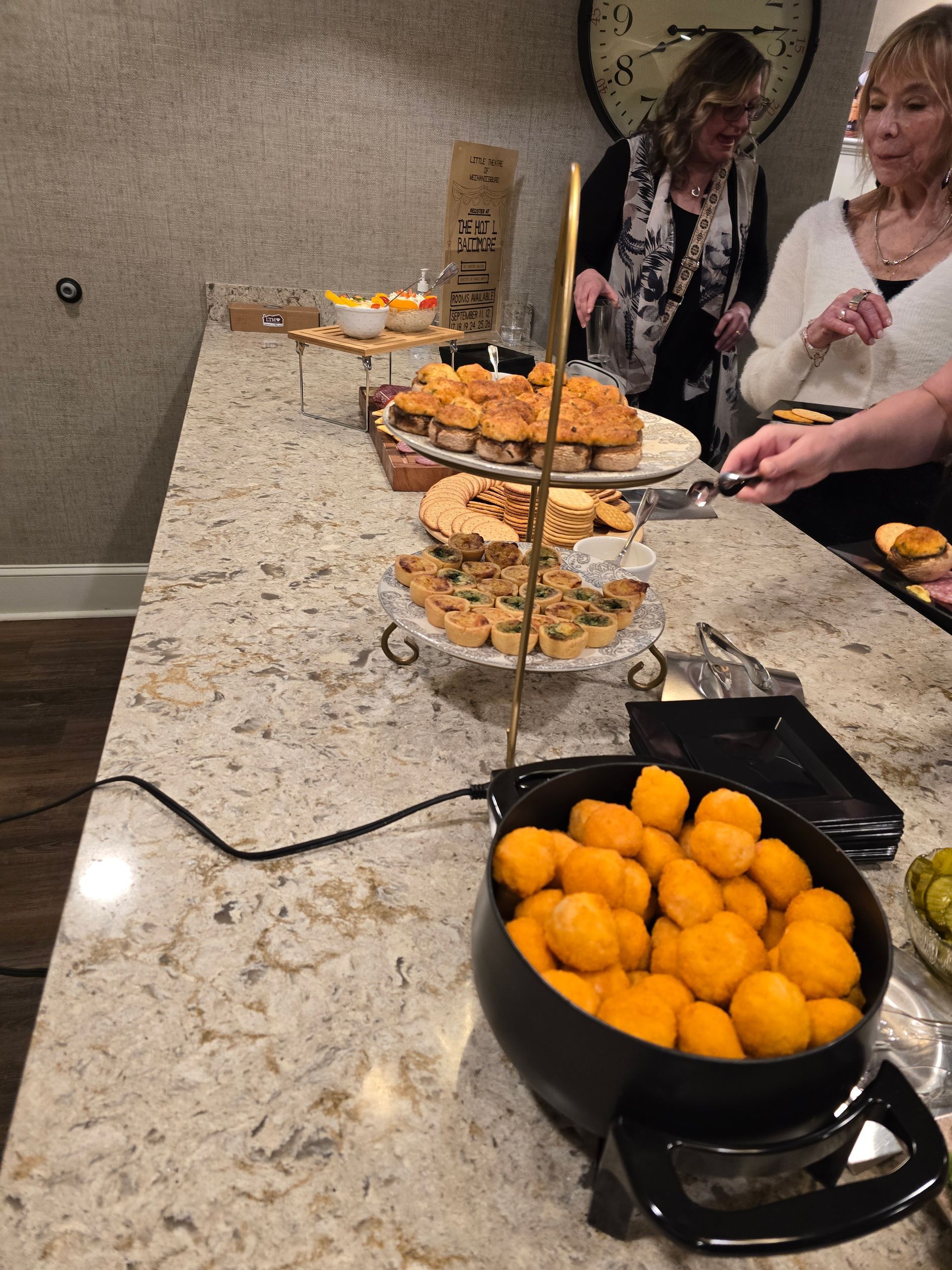 Buffet counter with trays of food and orange fried bites in a black skillet, with two people serving themselves.