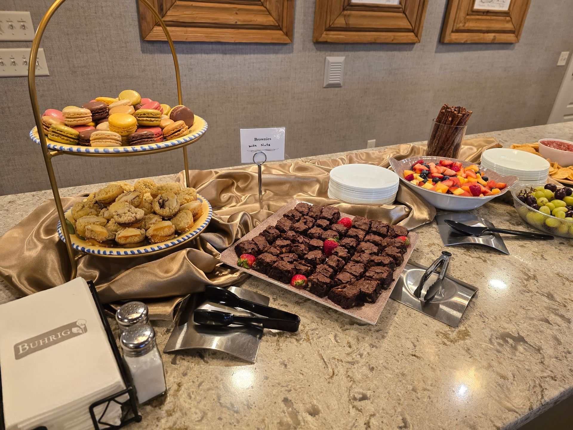 Dessert buffet with cupcakes, cookies, brownies, fruit bowls, and serving tongs on a marble counter