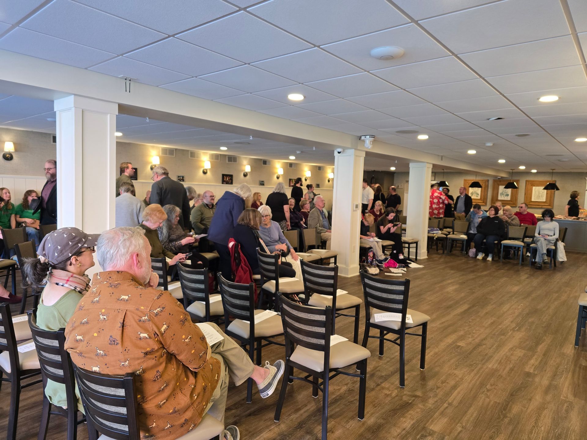 Indoor community gathering with seated audience in a bright hall, chairs arranged facing a side wall.