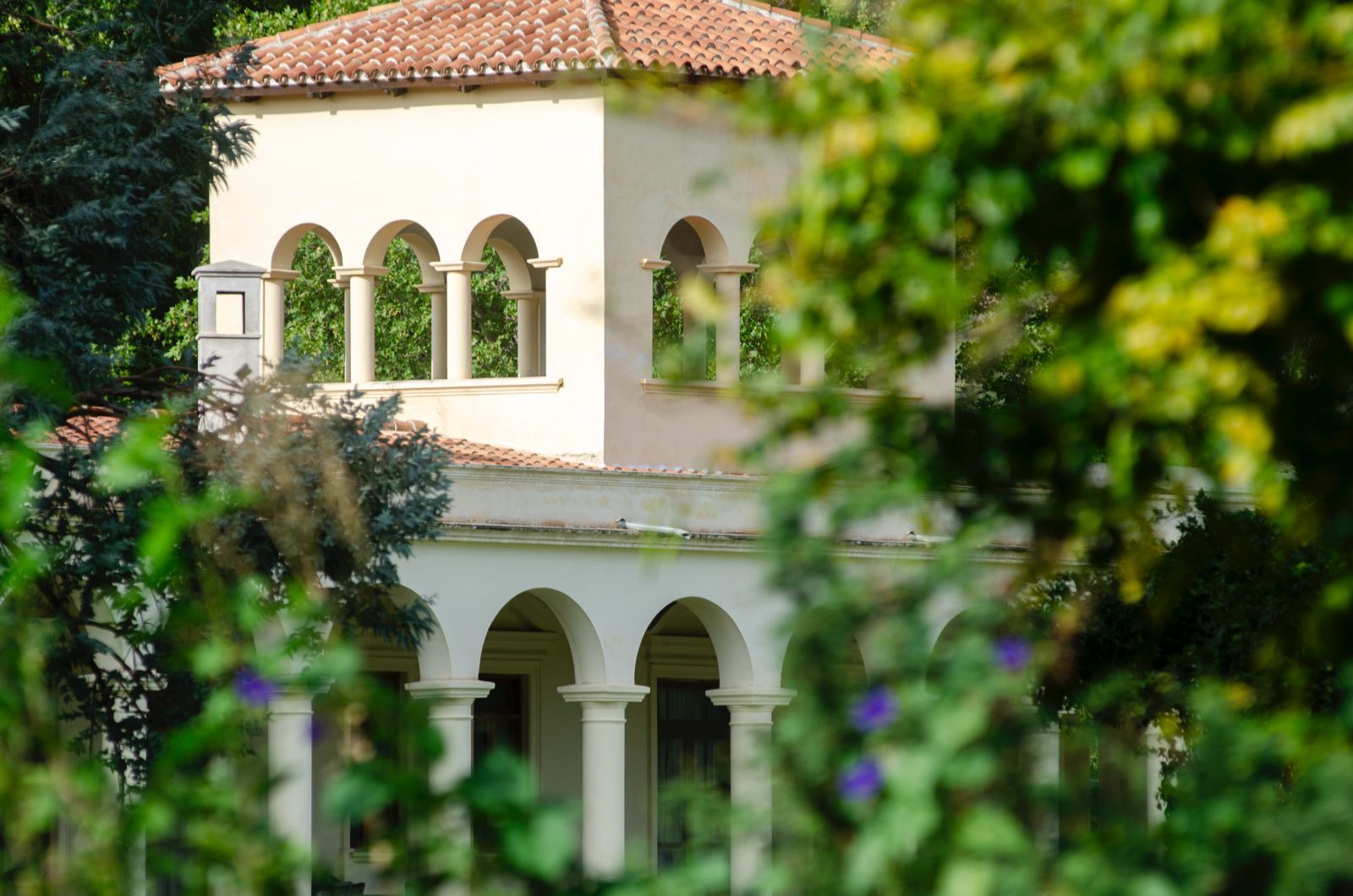 Beige building with arched windows and red tile roof, seen through green foliage.