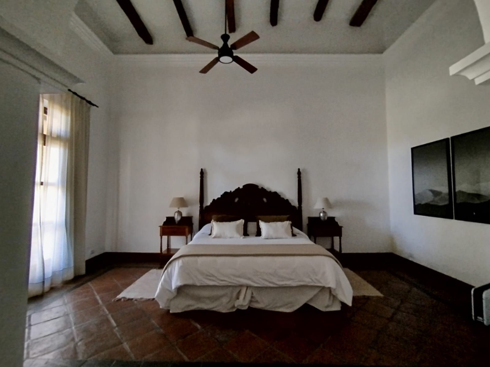 Bedroom with white walls, four-poster bed, wood beams, ceiling fan, and art.
