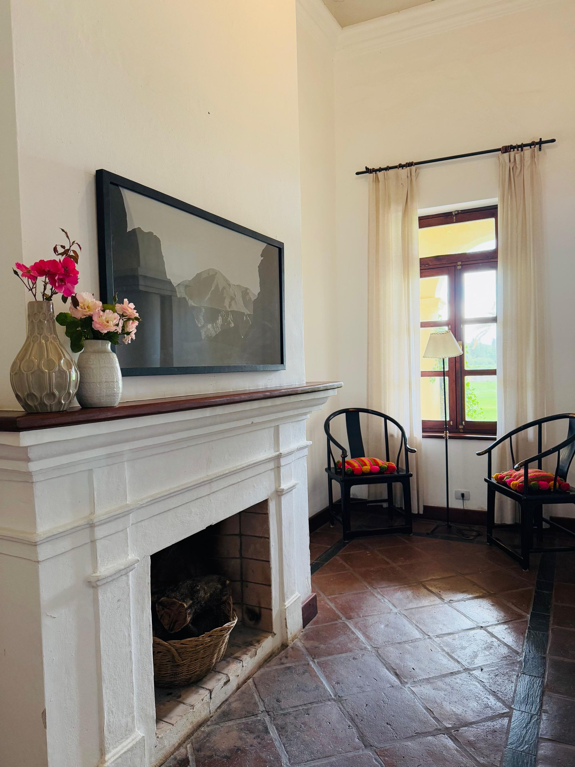 Fireplace with flowers, artwork, and chairs by a window in a white-walled room.