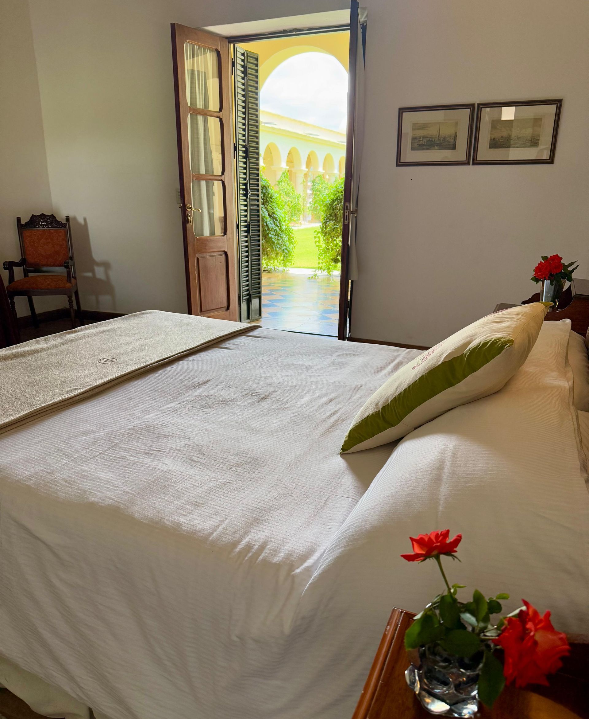 Bedroom with open door to a courtyard. White bedding, red roses, and a wooden chair.