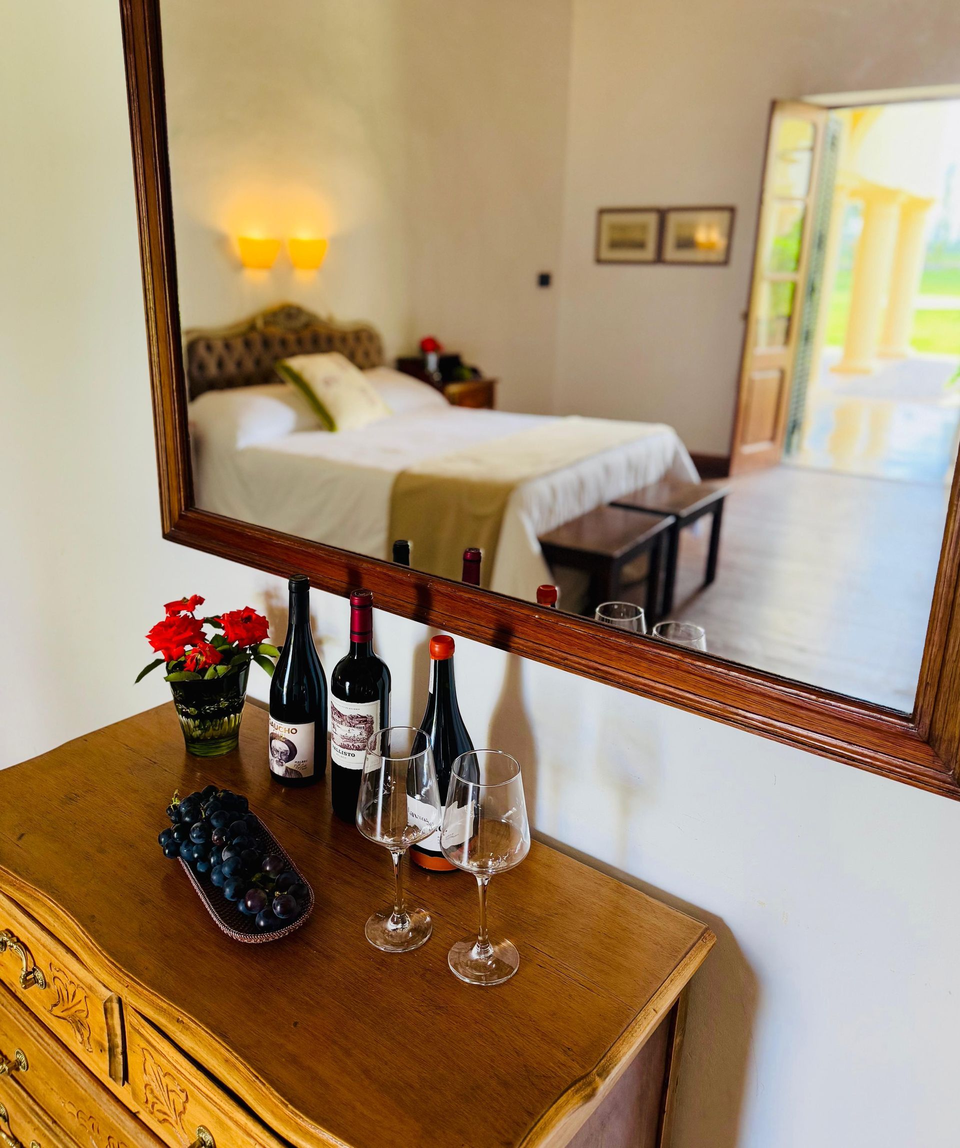 A bedroom reflected in a mirror. A table holds wine, glasses, and flowers.