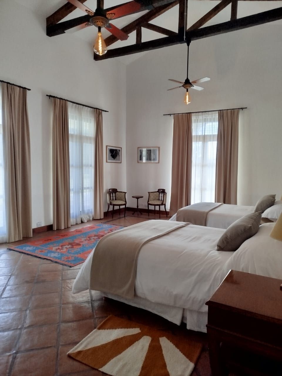 Bedroom with two beds, large windows, tan curtains, terracotta floors, area rugs, wood beams, and two ceiling fans.