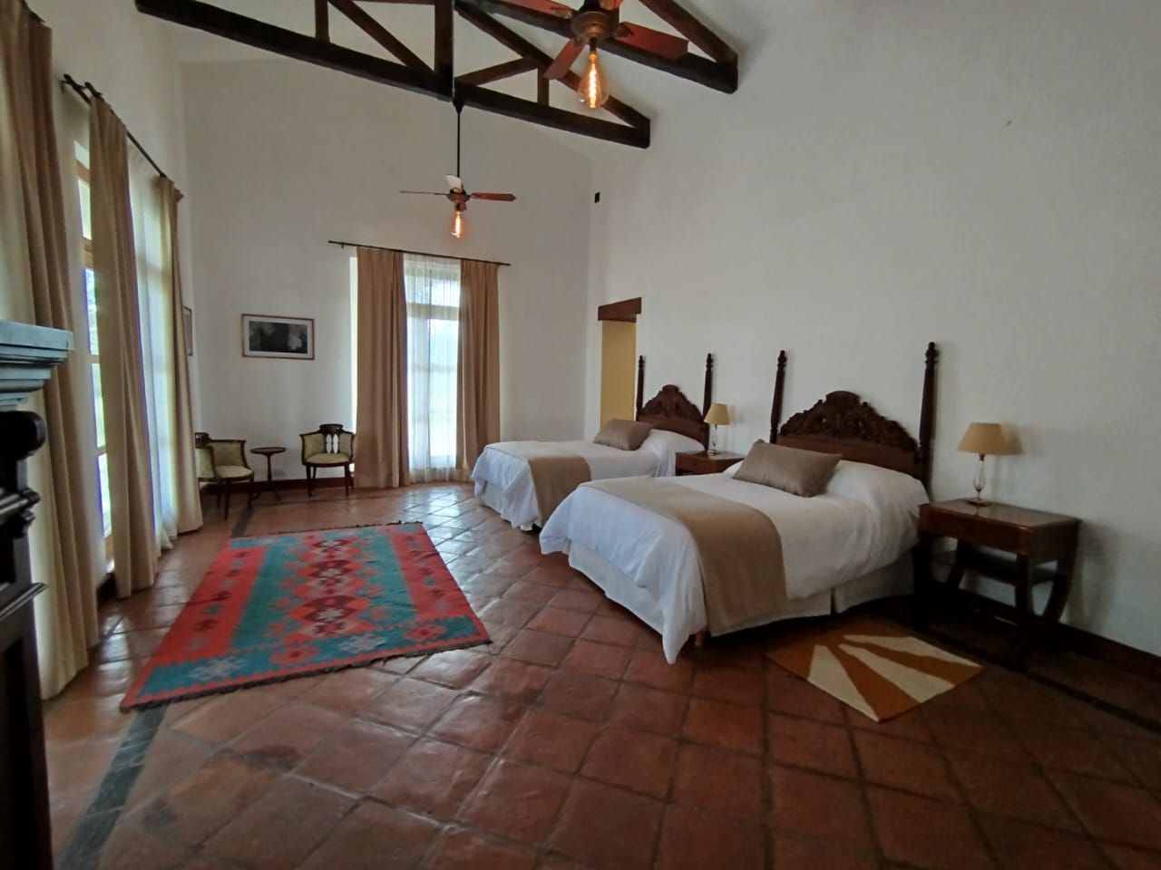 Bedroom with two beds, red rug, high ceiling, terracotta tile floor, and large windows with curtains.