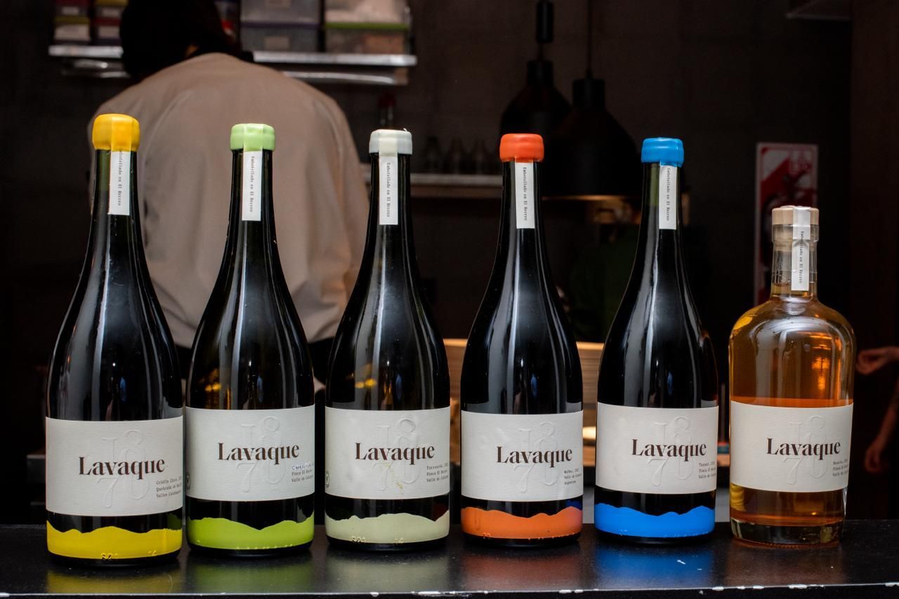 Six bottles of wine with colorful tops and bottoms on a bar, a person in the background.