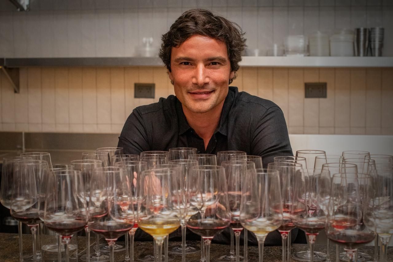 Man smiling behind a row of wine glasses filled with red and yellow liquid. Kitchen setting.