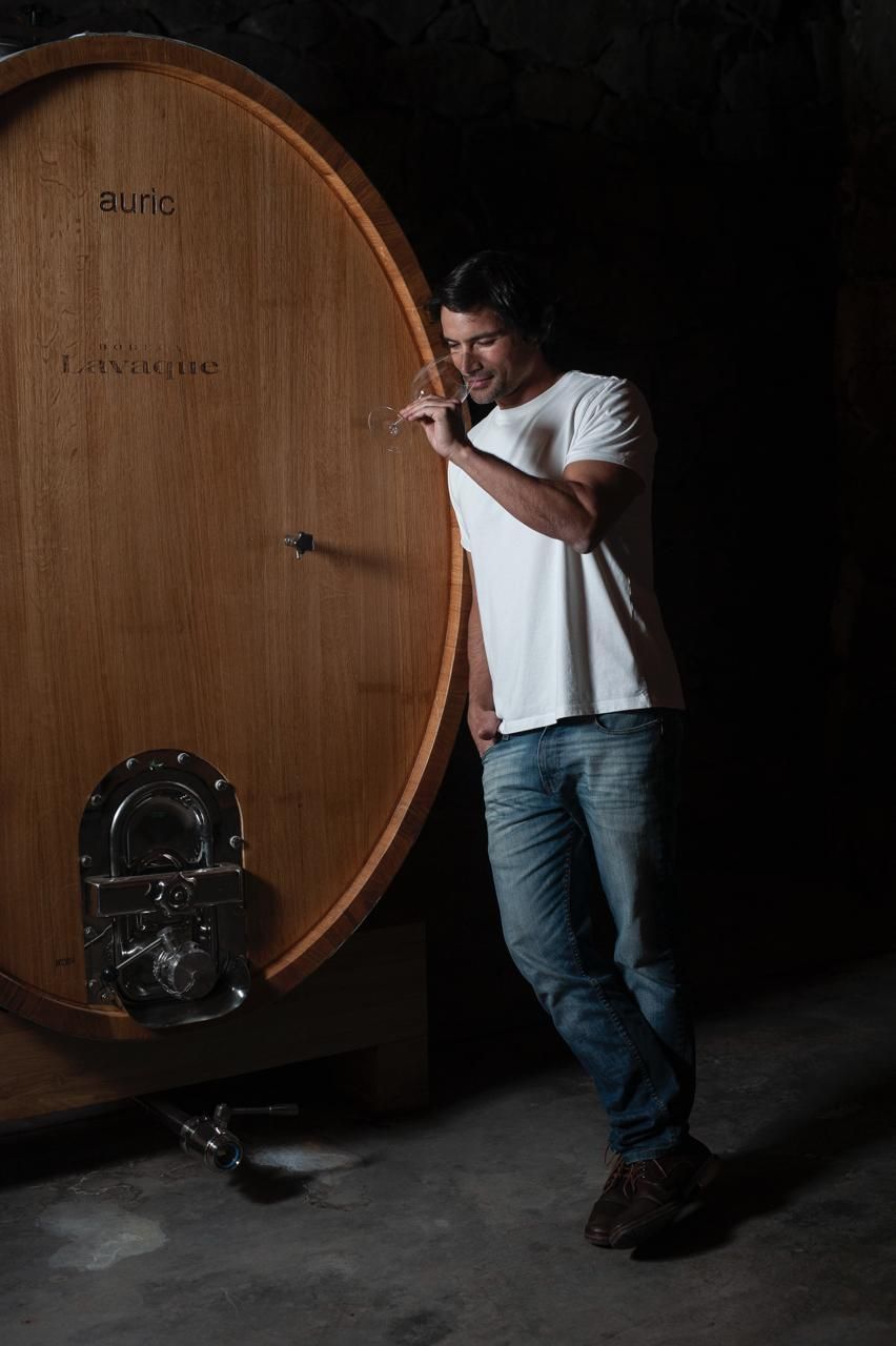 Man leaning against large wooden barrel in a dark cellar, smiling.