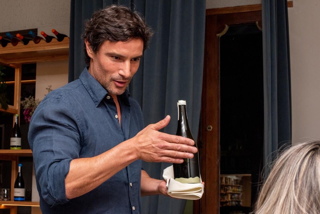 Man in blue shirt presenting a wine bottle indoors, gesturing.