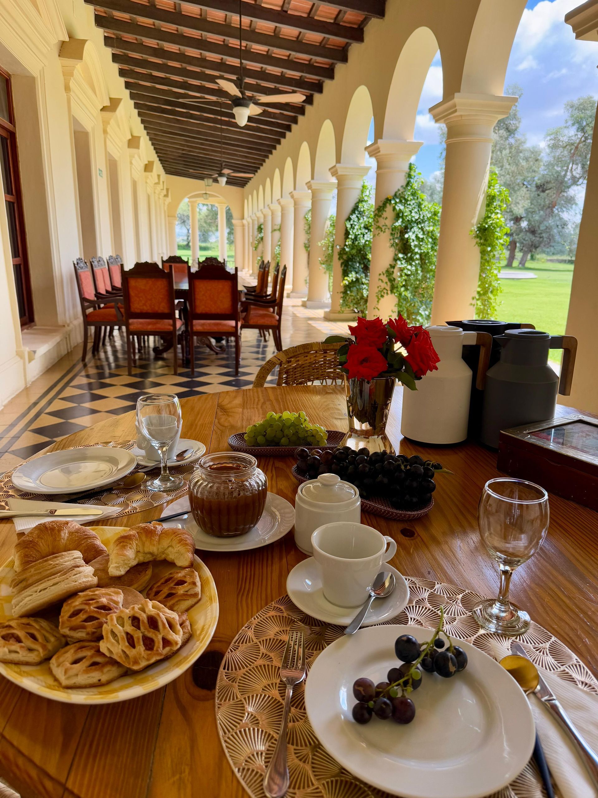 Breakfast table set on a veranda with pastries, fruit, and coffee. Overlooks a grassy lawn.