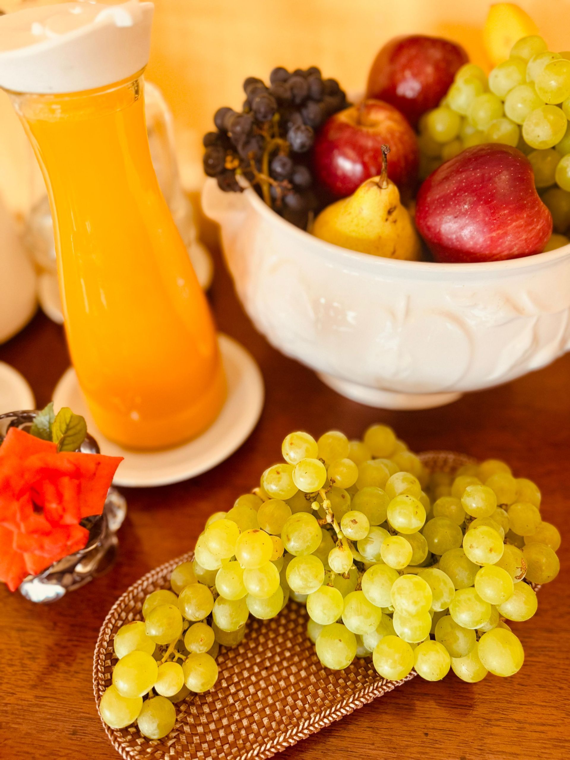 A fruit bowl of apples, grapes, and pear sits beside a juice pitcher and a plate with watermelon.