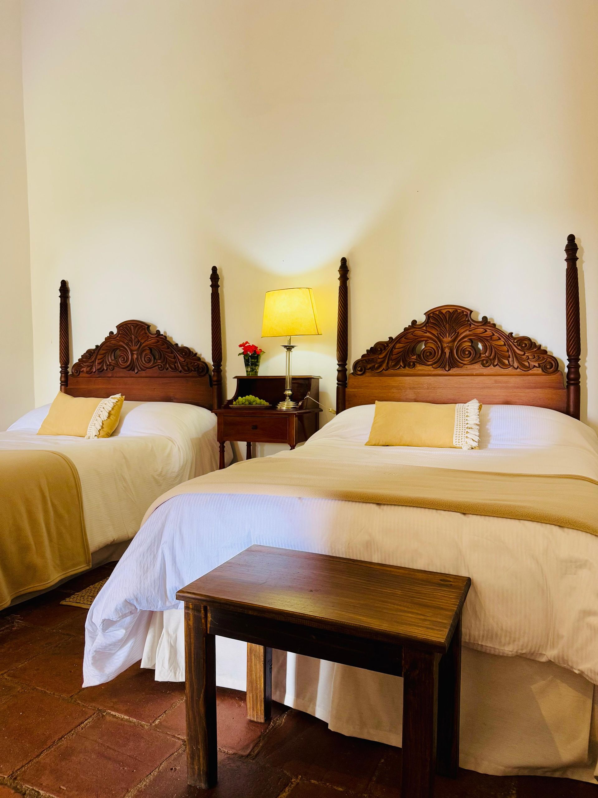 Bedroom with two beds, ornate wooden headboards, nightstand with lamp, and a small wooden bench.