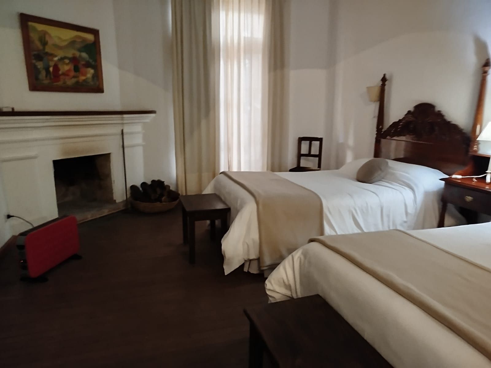 Bedroom with two beds, fireplace, small table, and a window with sheer curtains.