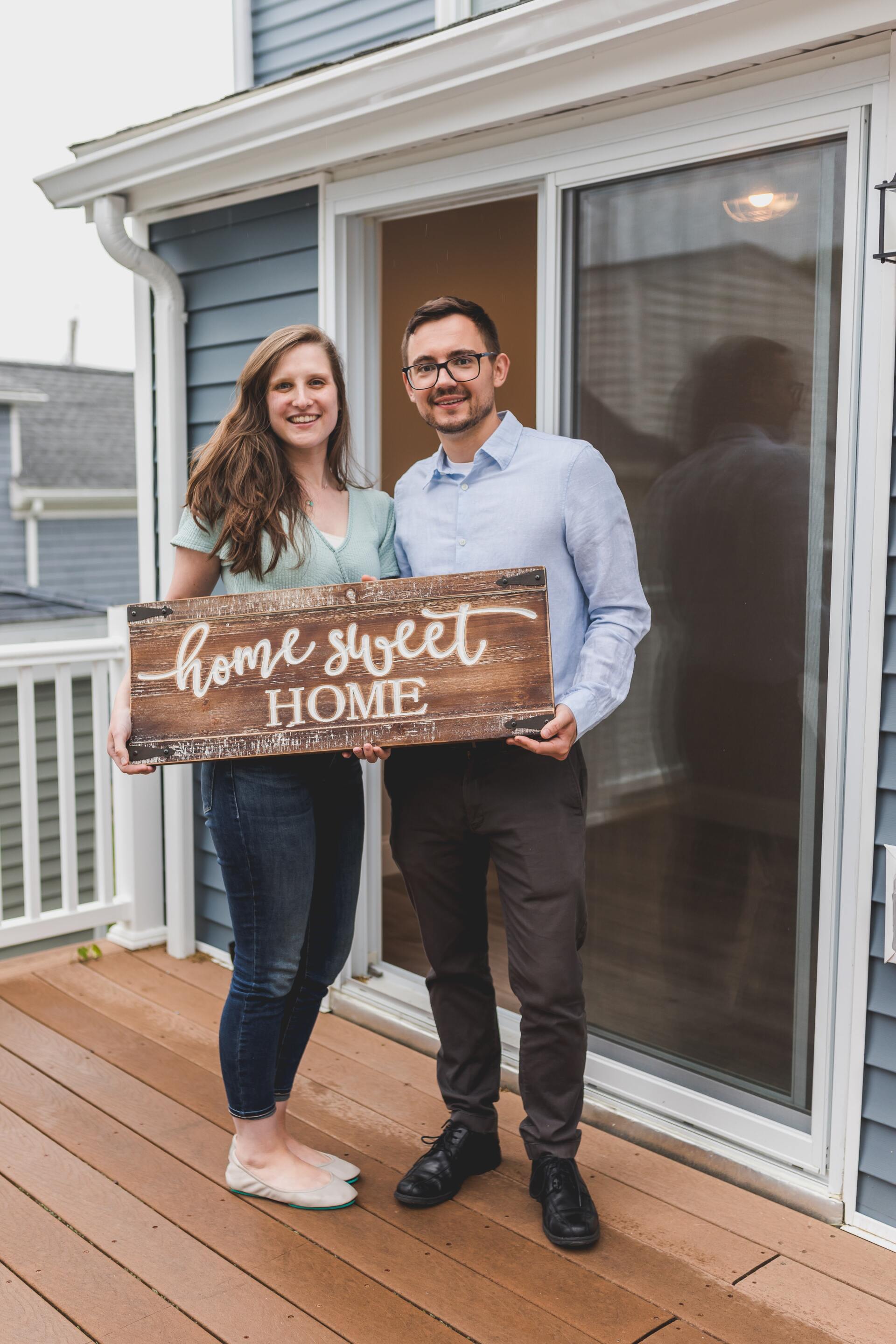 Couple standing on a deck holding a “home sweet home” sign by a sliding glass door