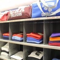 Folded sports jerseys stacked on shelves, with red, blue, and purple sports bags above.