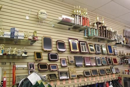 Wall of assorted trophies and framed plaques displayed on slatwall shelves in a store.