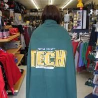 Person wrapped in a teal TECH blanket in a clothing store aisle, seen from behind.