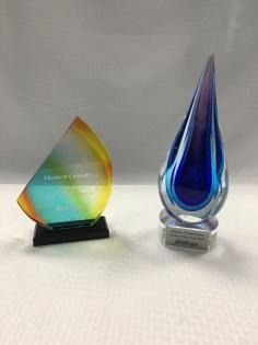 Two colorful glass awards on a white surface, one flame-shaped blue and the other gold-green.