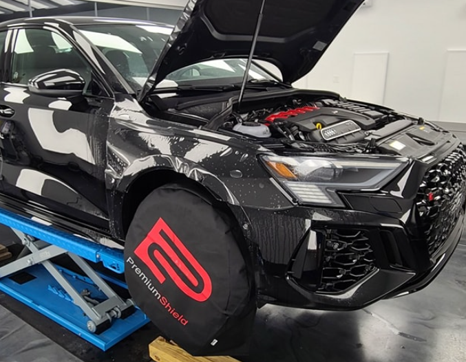 A black car with the hood up is sitting on a lift.