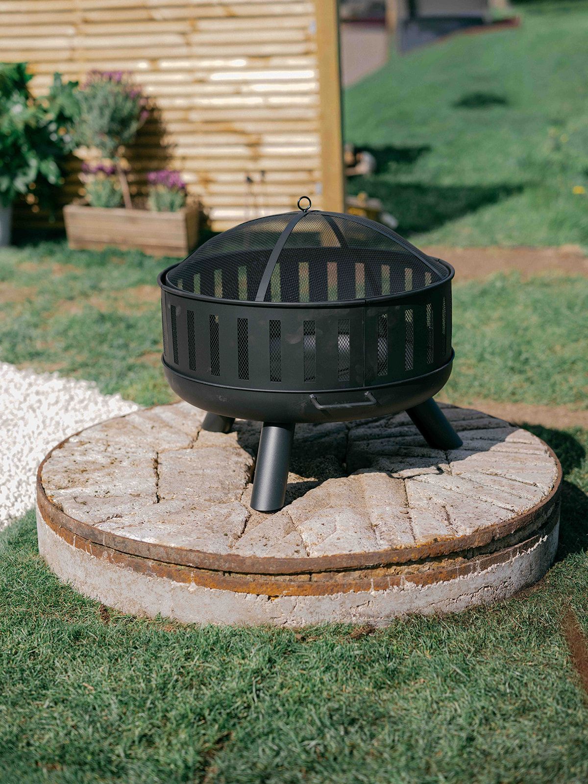 Black metal fire pit on a round stone base in a grassy yard.