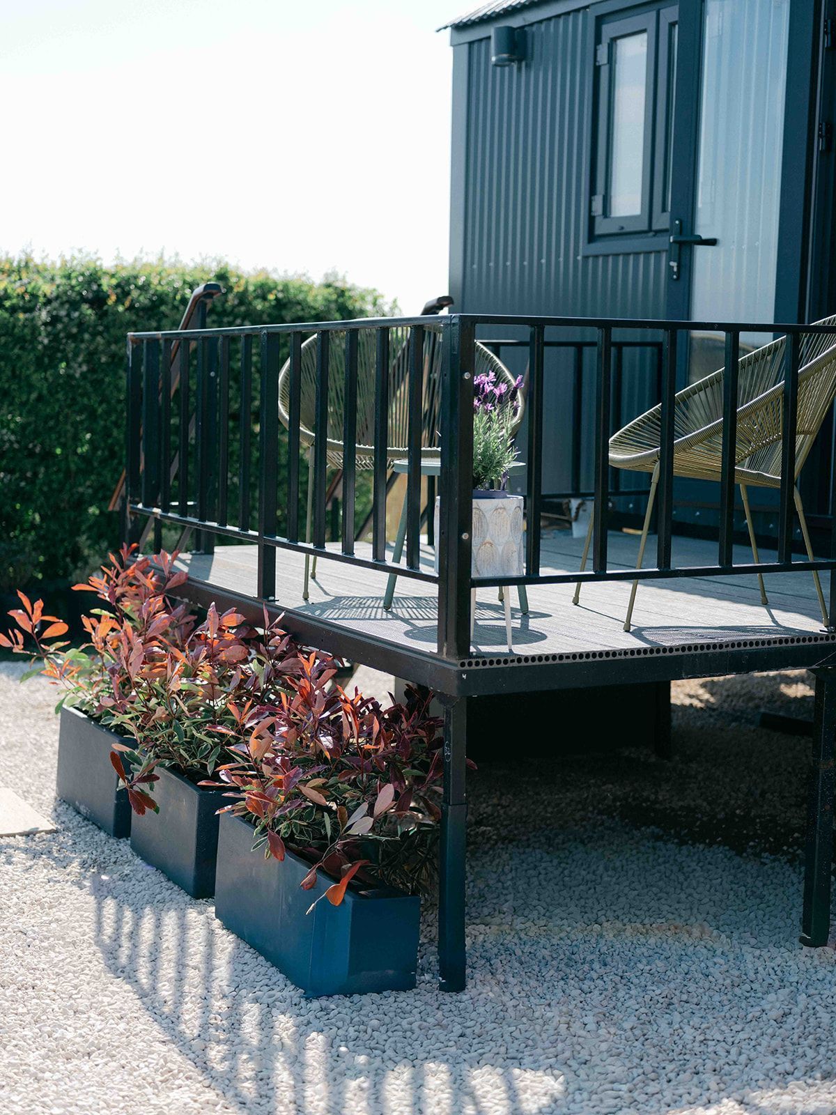 Black metal patio deck beside a modern cabin, with potted red plants and outdoor chairs