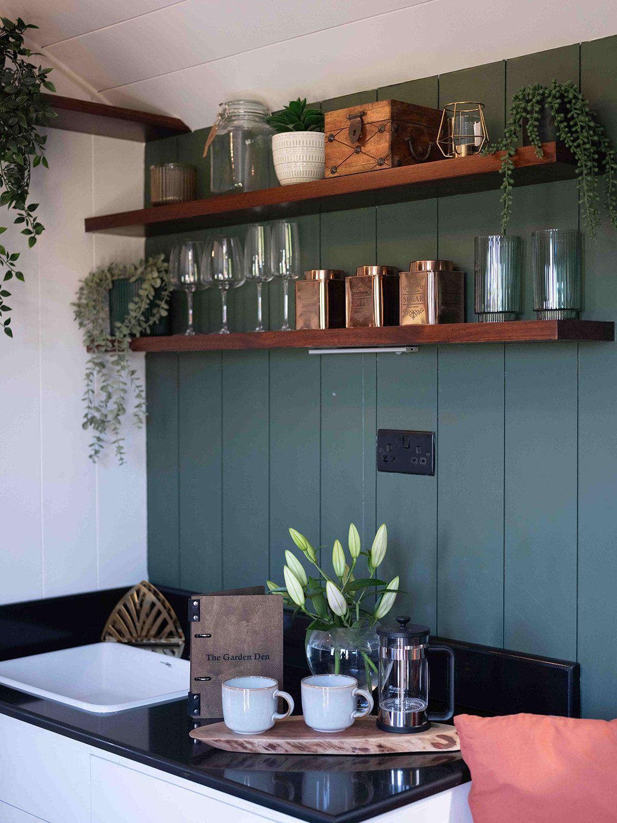 Cozy kitchen nook with green paneled wall, shelves, glass jars, mugs, flowers, and a black table with white cups