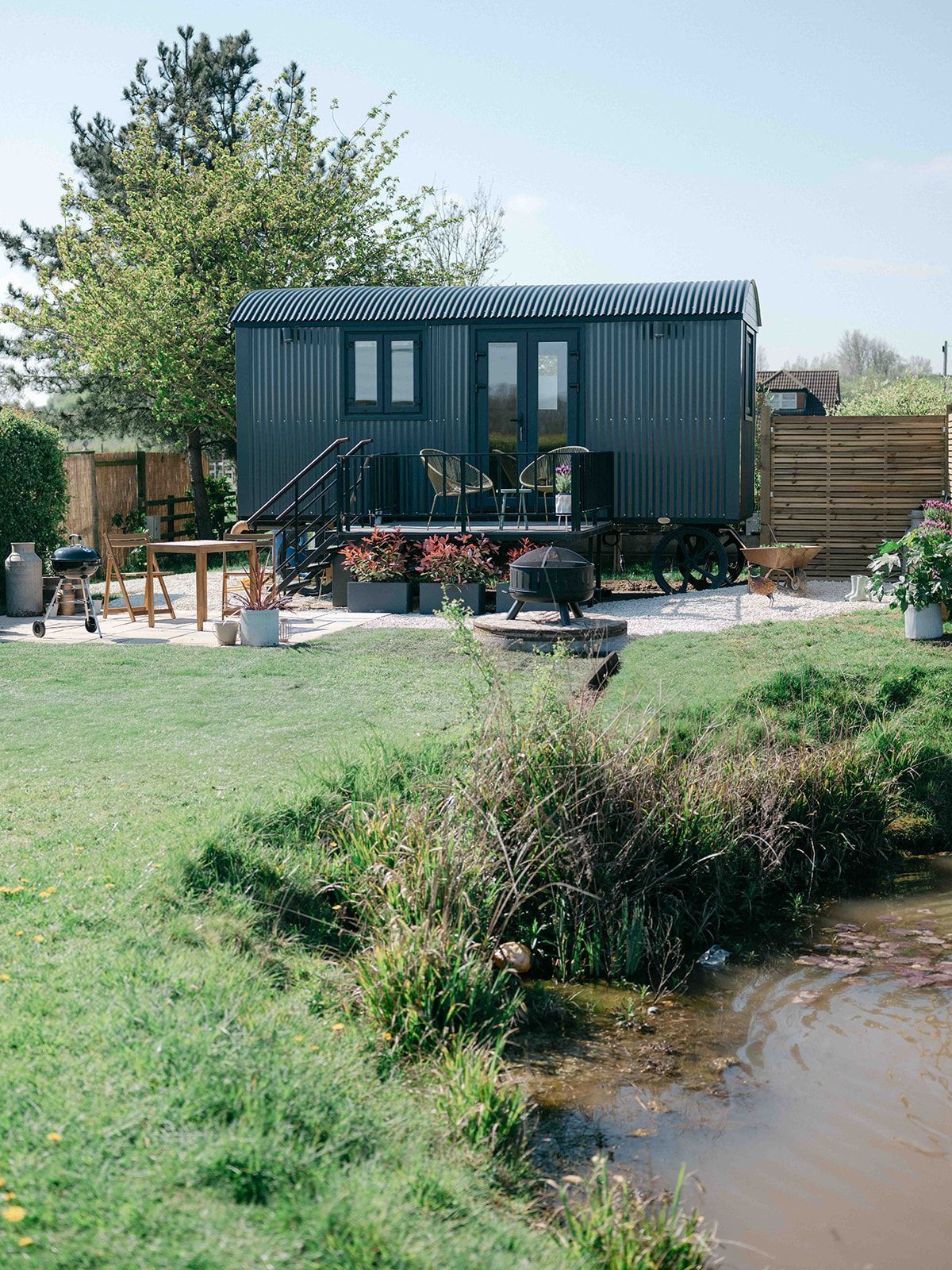 Dark green cabin on a grassy bank beside a pond, with trees and a patio table outside.