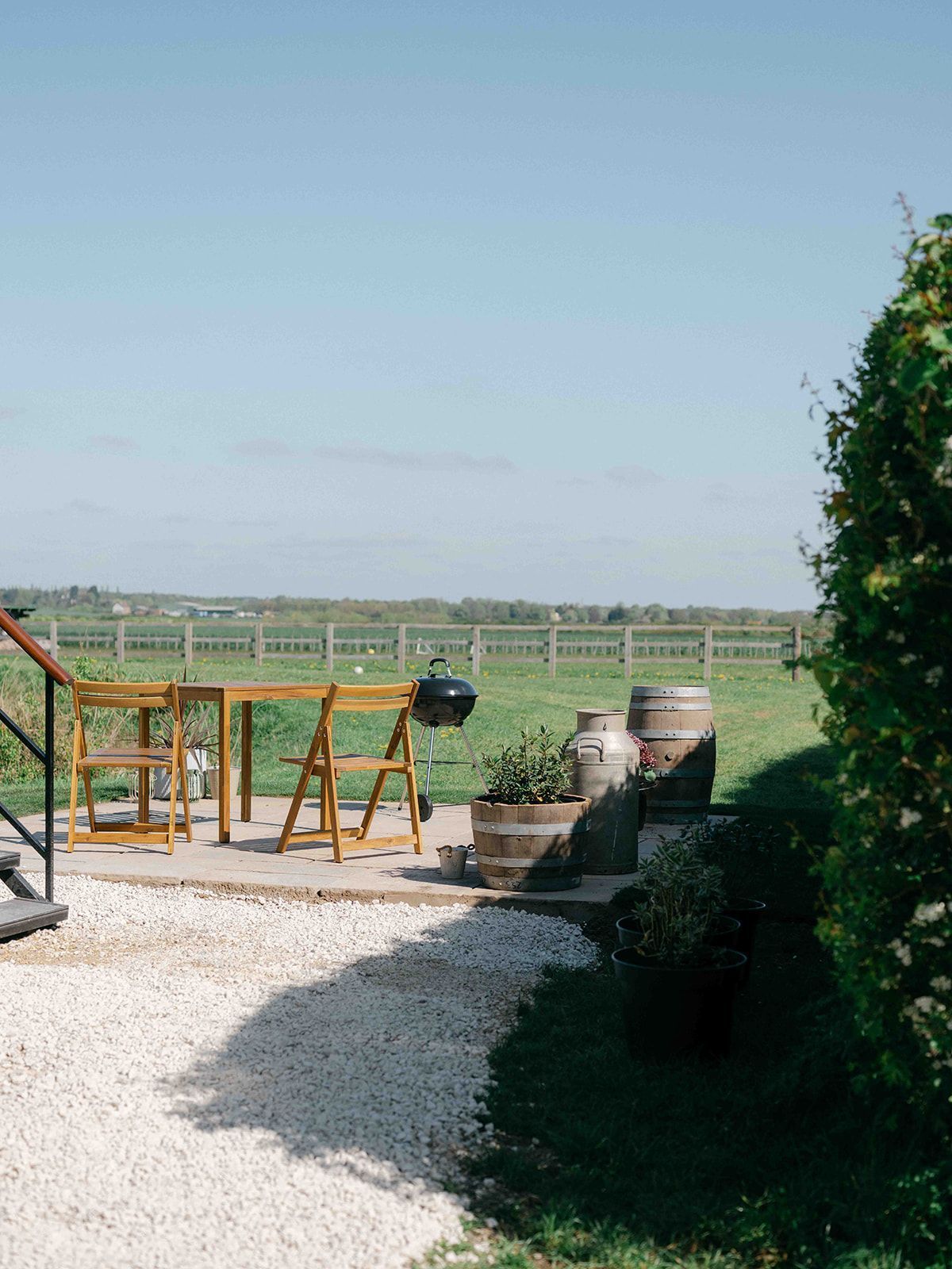 Patio with wooden table, grill, and potted plants overlooking a green countryside field