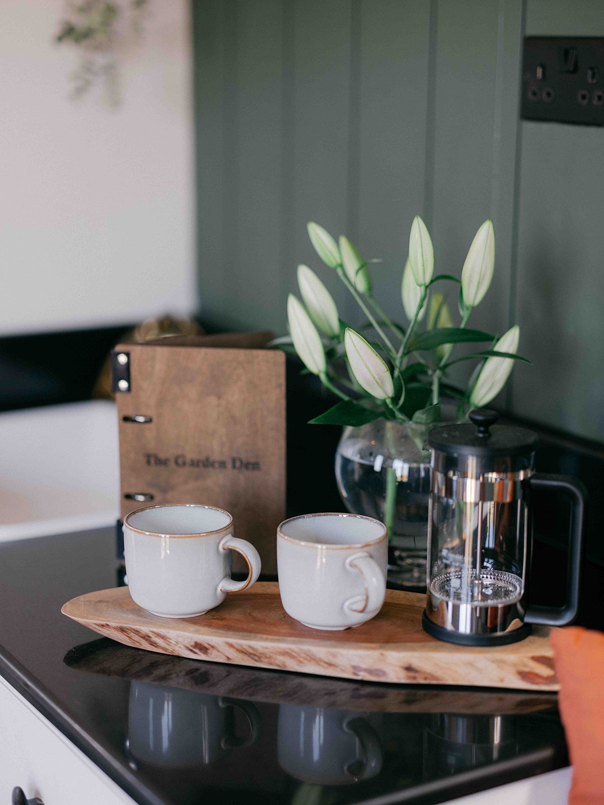 Two white mugs on a wooden tray beside a glass vase with white flowers on a black countertop