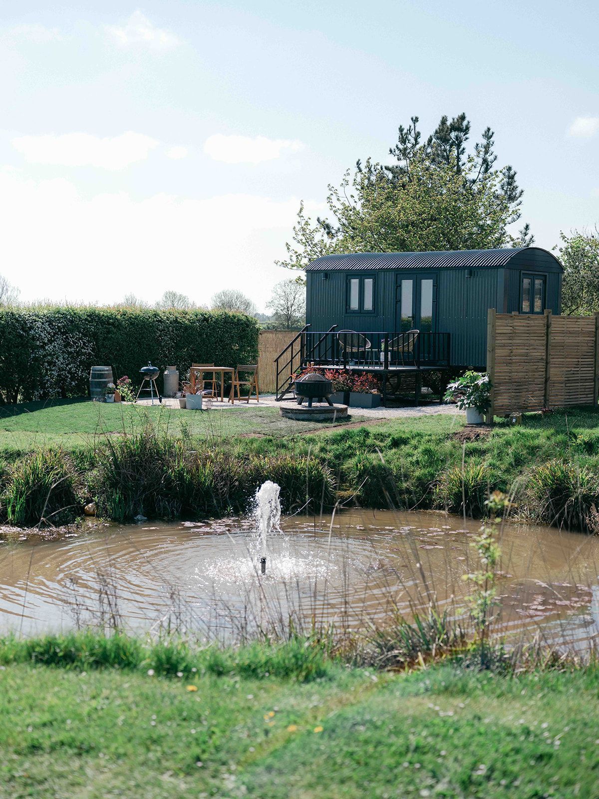 Small pond with a fountain in a grassy garden beside a dark shed and patio seating