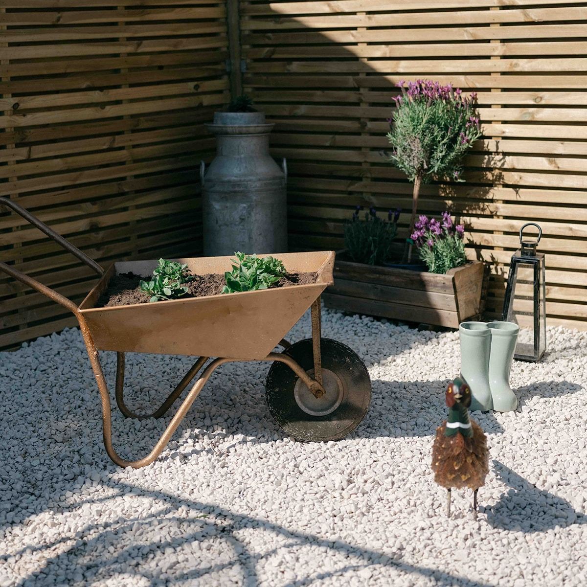 Rustic patio with a wheelbarrow planter, potted flowers, lanterns, and gravel against a wooden fence.