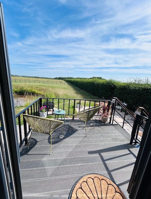 Patio deck with black railing, wicker chairs, and fields under a blue sky beyond the hedge