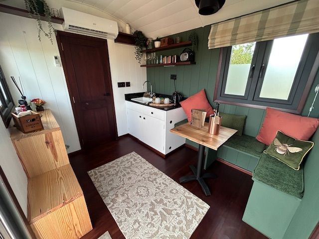 Cozy tiny room with green bench, small table, sink, shelves, and rug beside a wooden door and window.