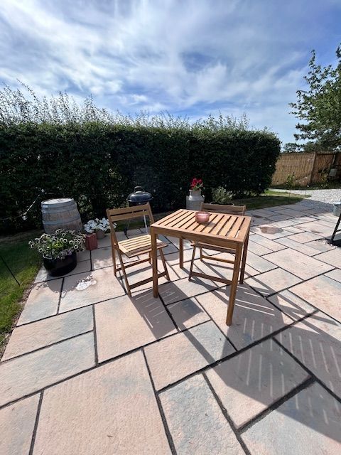 Patio with wooden table and chairs on a sunlit stone terrace, bordered by hedges and potted plants.
