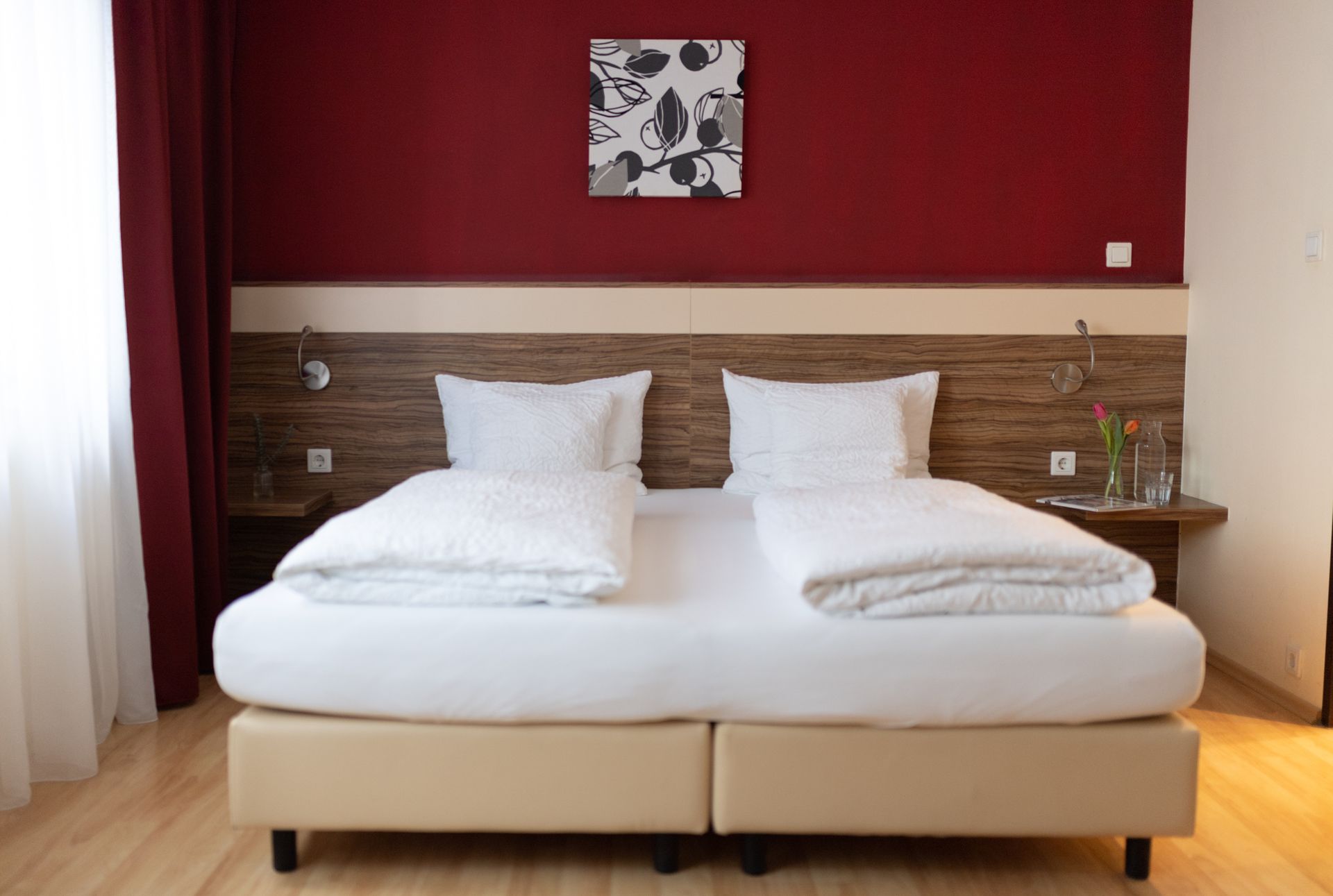 Großes Doppelbett. Das Bett steht an einer Weinroten Wand und neben dem Bett sind je ein Nachttisch.