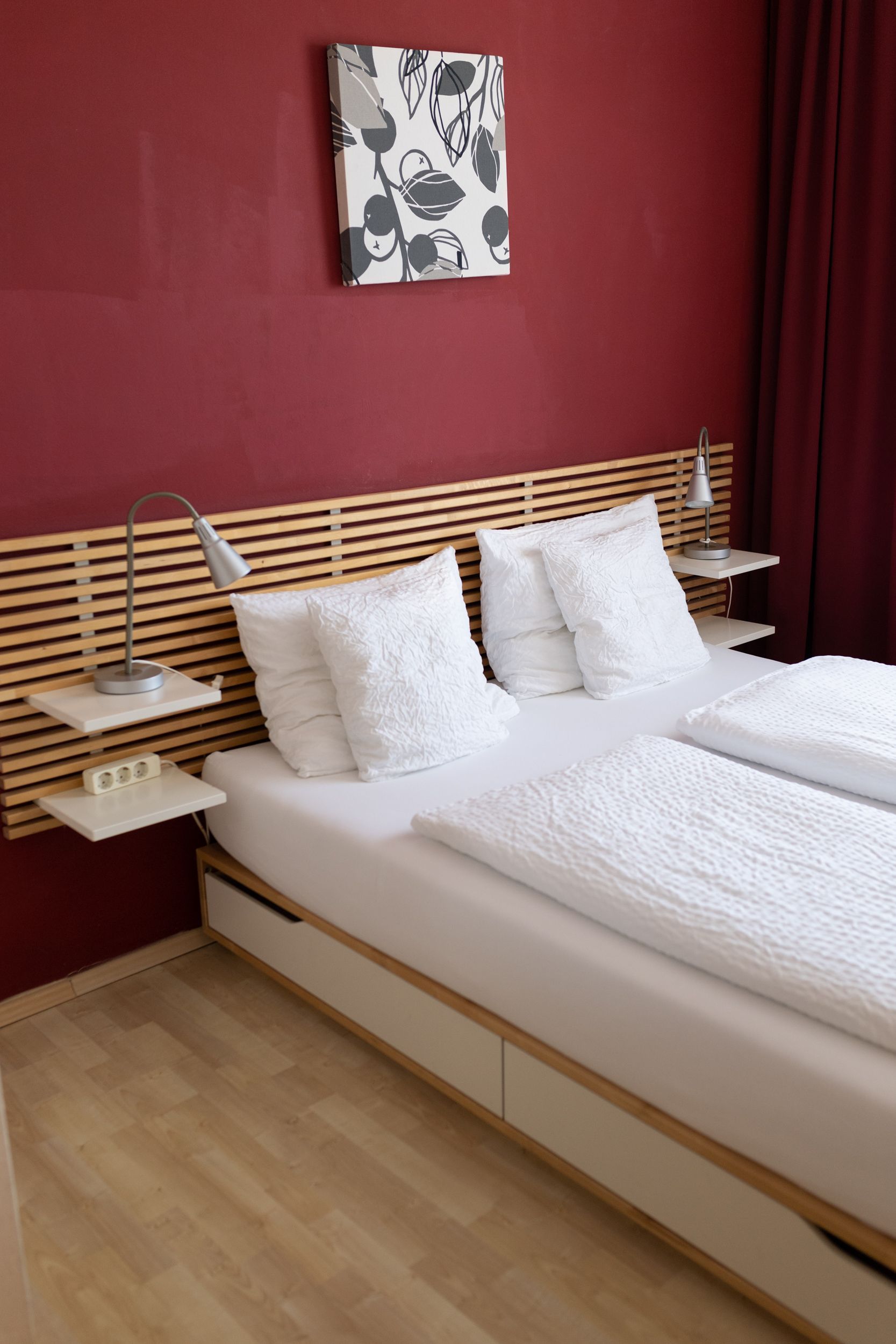 Großes Doppelbett. Das Bett steht an einer Weinroten Wand und neben dem Bett sind je ein Nachttisch.