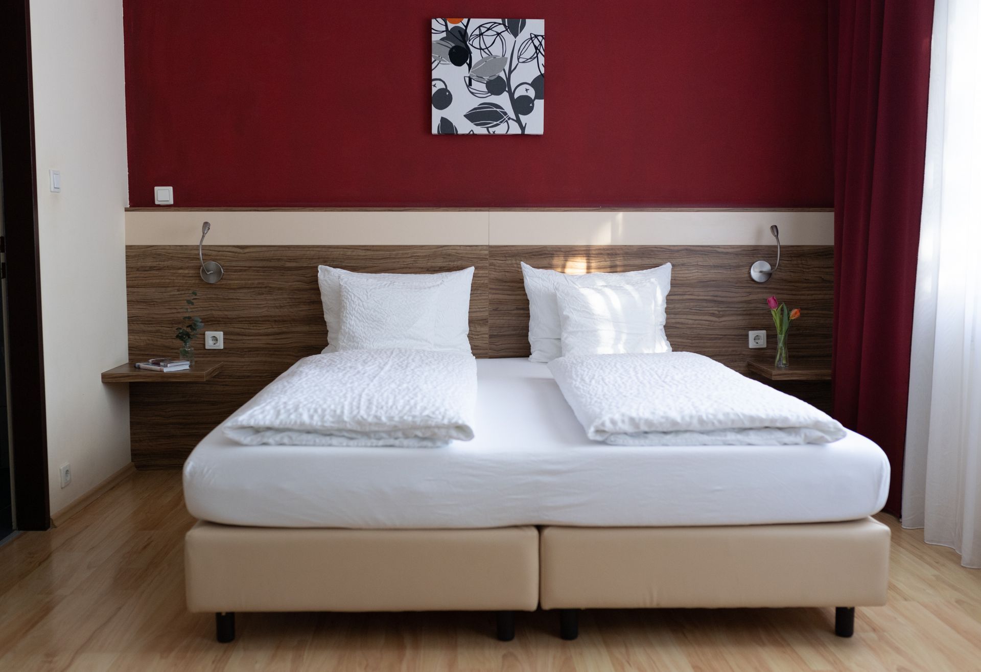Großes Doppelbett. Das Bett steht an einer Weinroten Wand und neben dem Bett sind je ein Nachttisch.