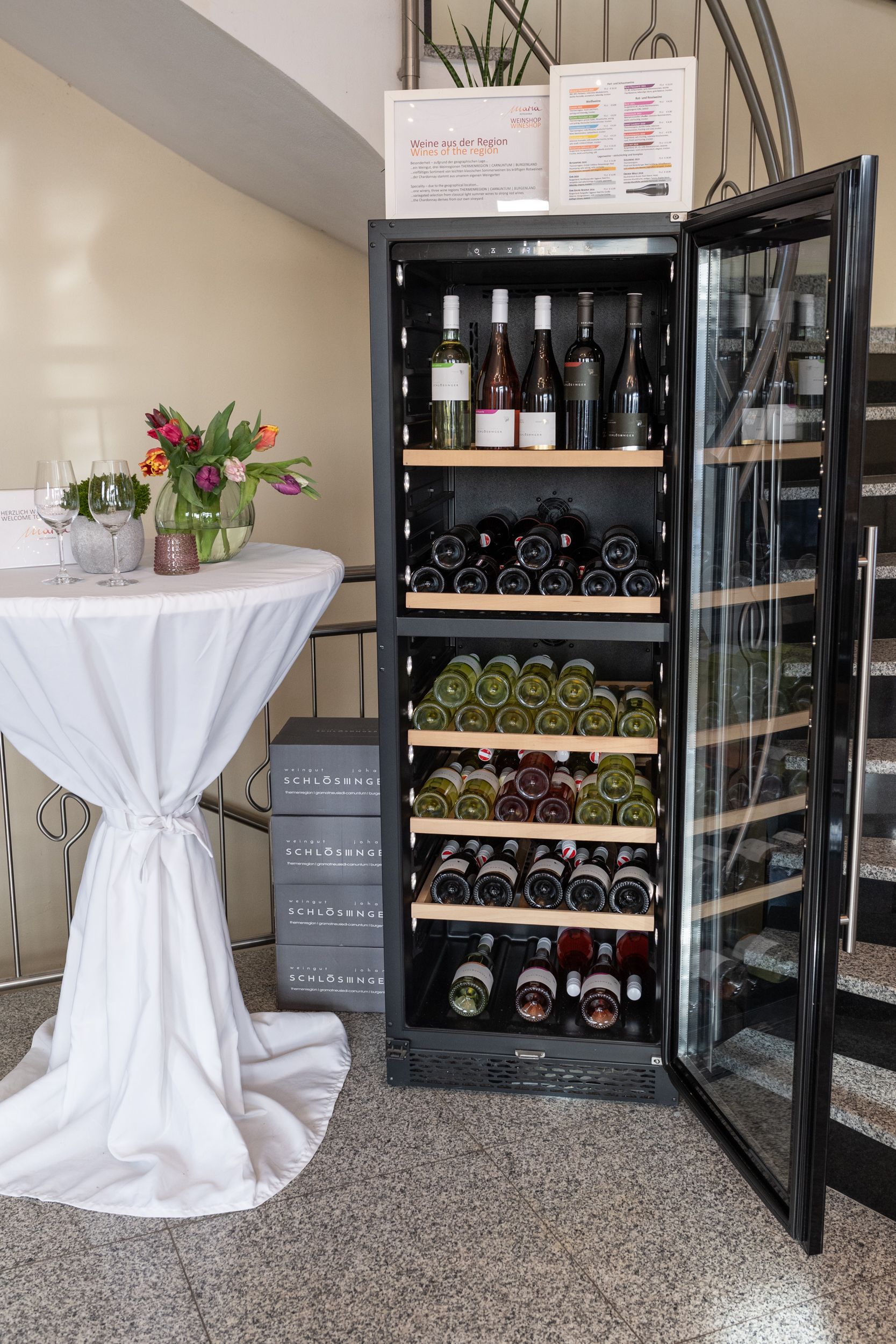 Empfang HotelMaria. Geöffneter Weinkühlschrank mit lokalen Weinen. Daneben ein Tisch mit Weingläsern und bunten Tulpen.