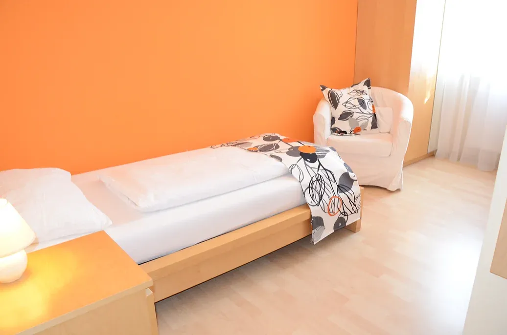 Einzelzimmer mit Einzelbett, warm eingerichtet. Das Bett steht an einer orange gestrichenen Wand