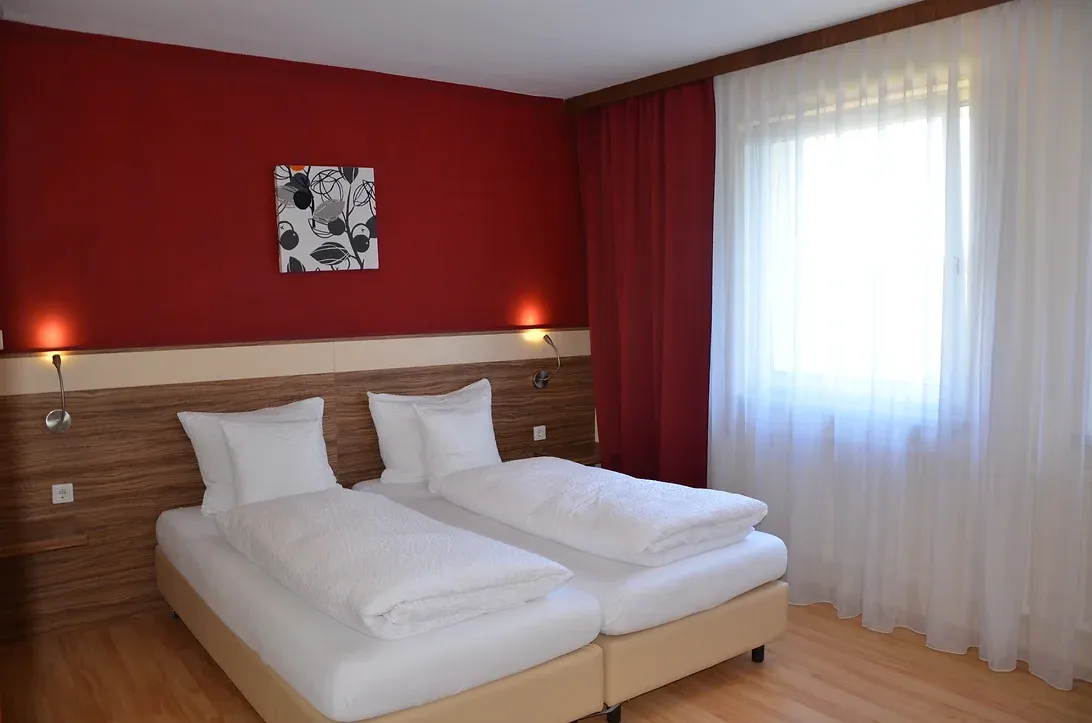 Großes Doppelbett. Das Bett steht an einer Weinroten Wand und neben dem Bett sind je ein Nachttisch.