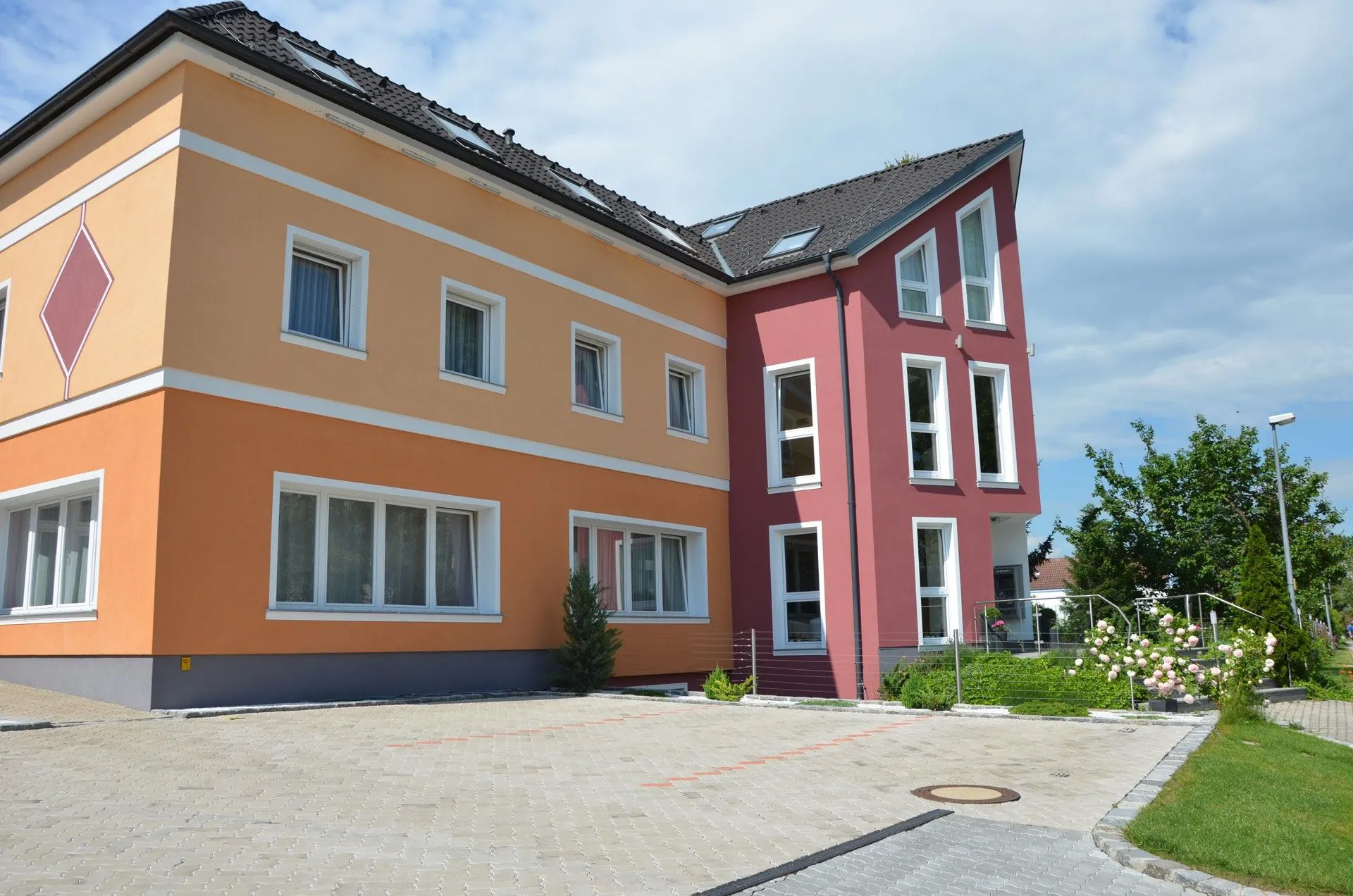 HotelMaria Außenbereich. Rot und orangene Fassade. Großer Parkplatz mit Pflastersteinbelag.