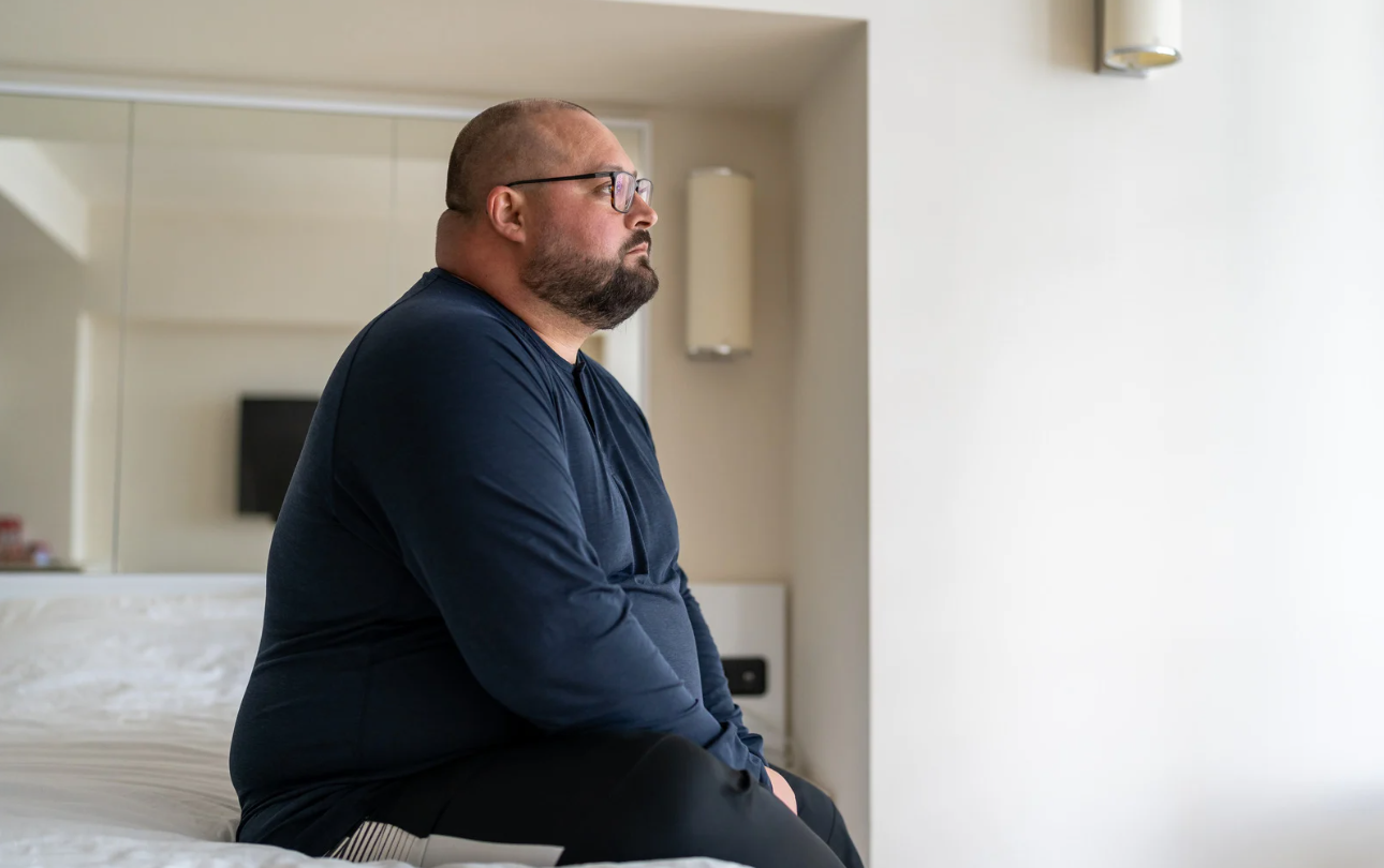 Man sitting on a bed in a bright room, looking out a window.