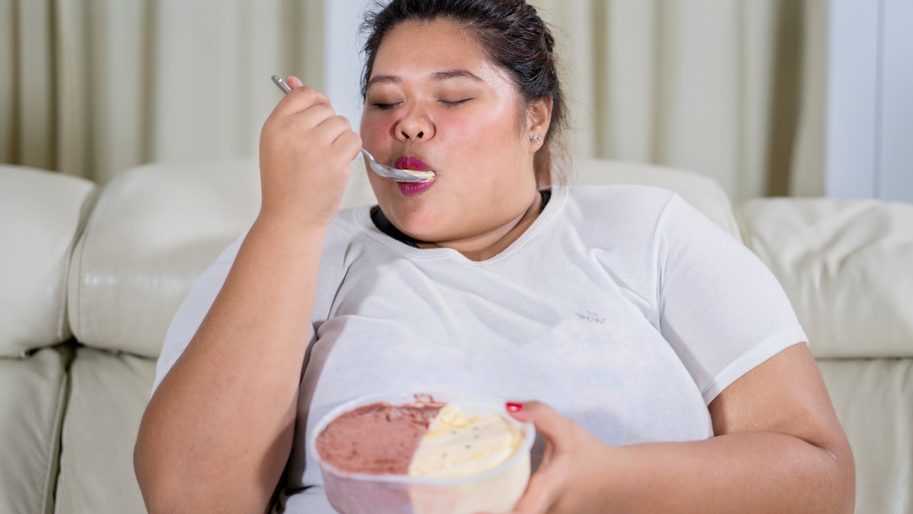 Woman eating ice cream on a couch, wearing a white shirt and holding a bowl in a bright living room.