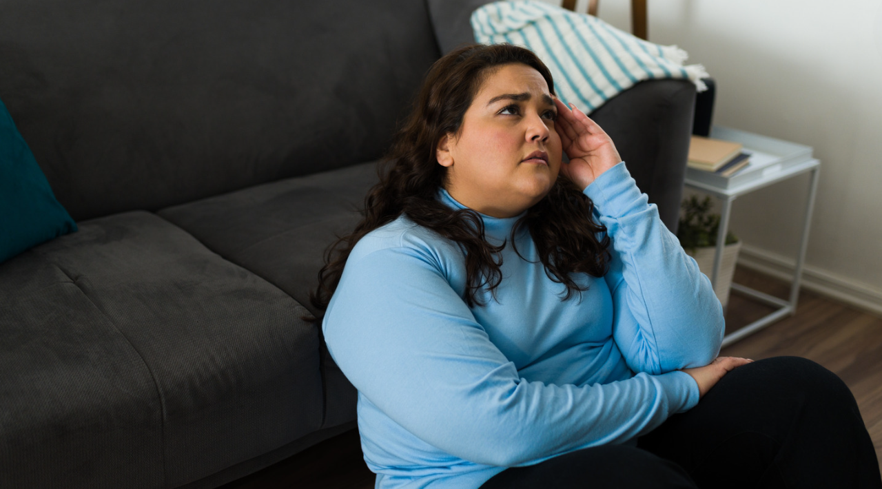 Person in a blue sweater sits on a couch, resting a hand on their head and looking concerned.