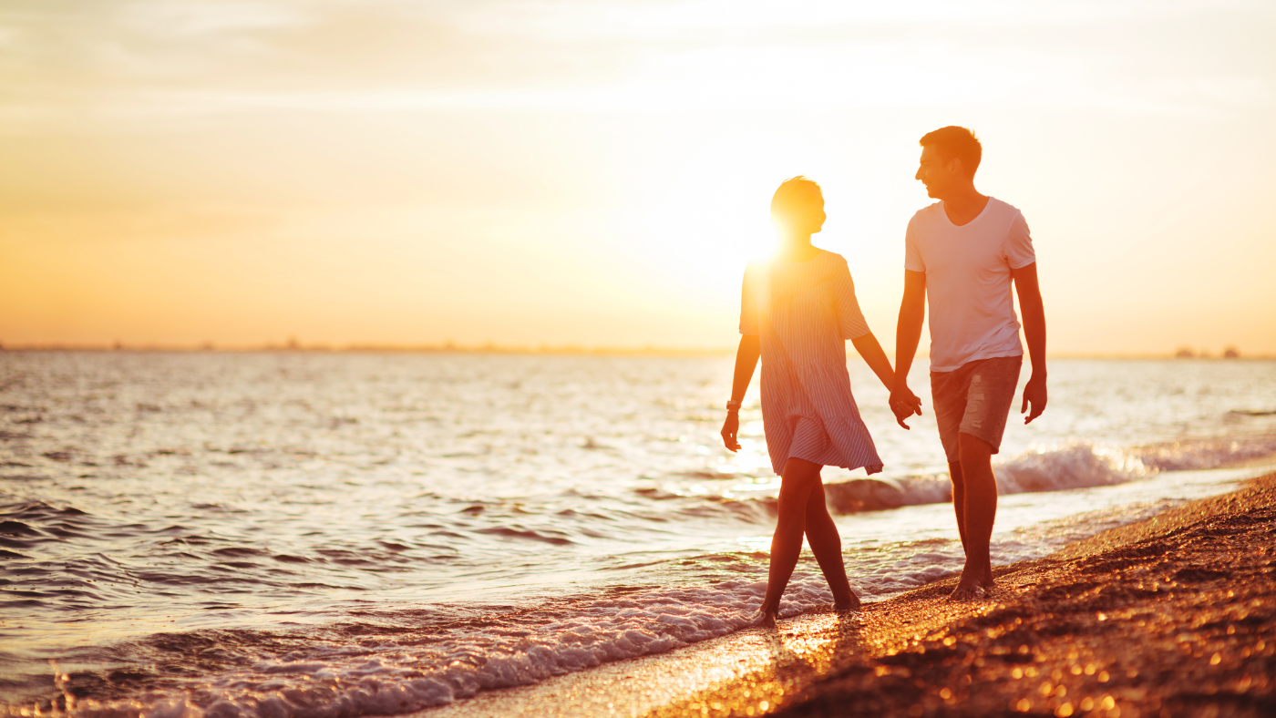 Two people holding hands walking along a beach at sunset, with warm golden light on the water and shore