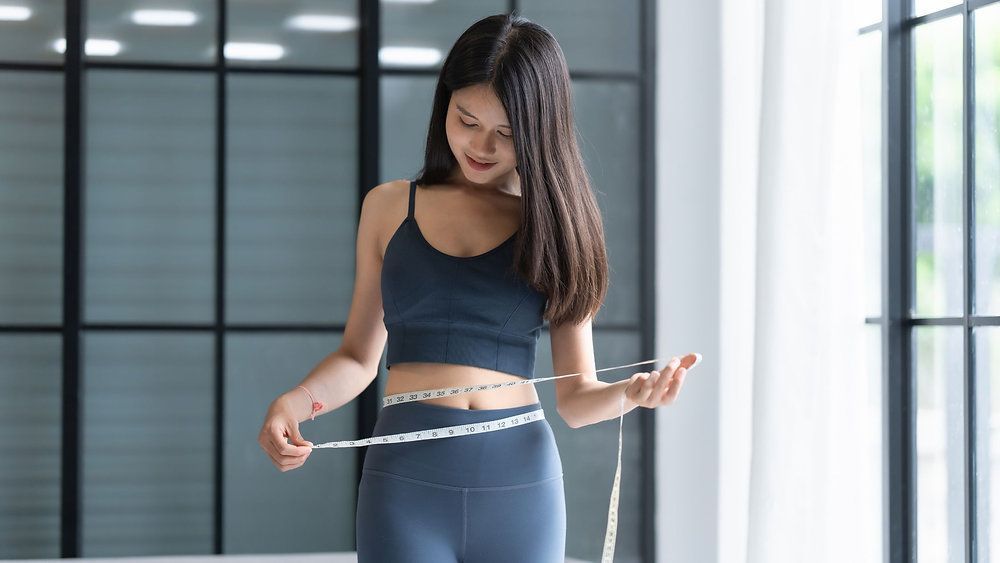 Mulher com roupa de ginástica medindo a cintura com uma fita métrica em ambiente interno.
