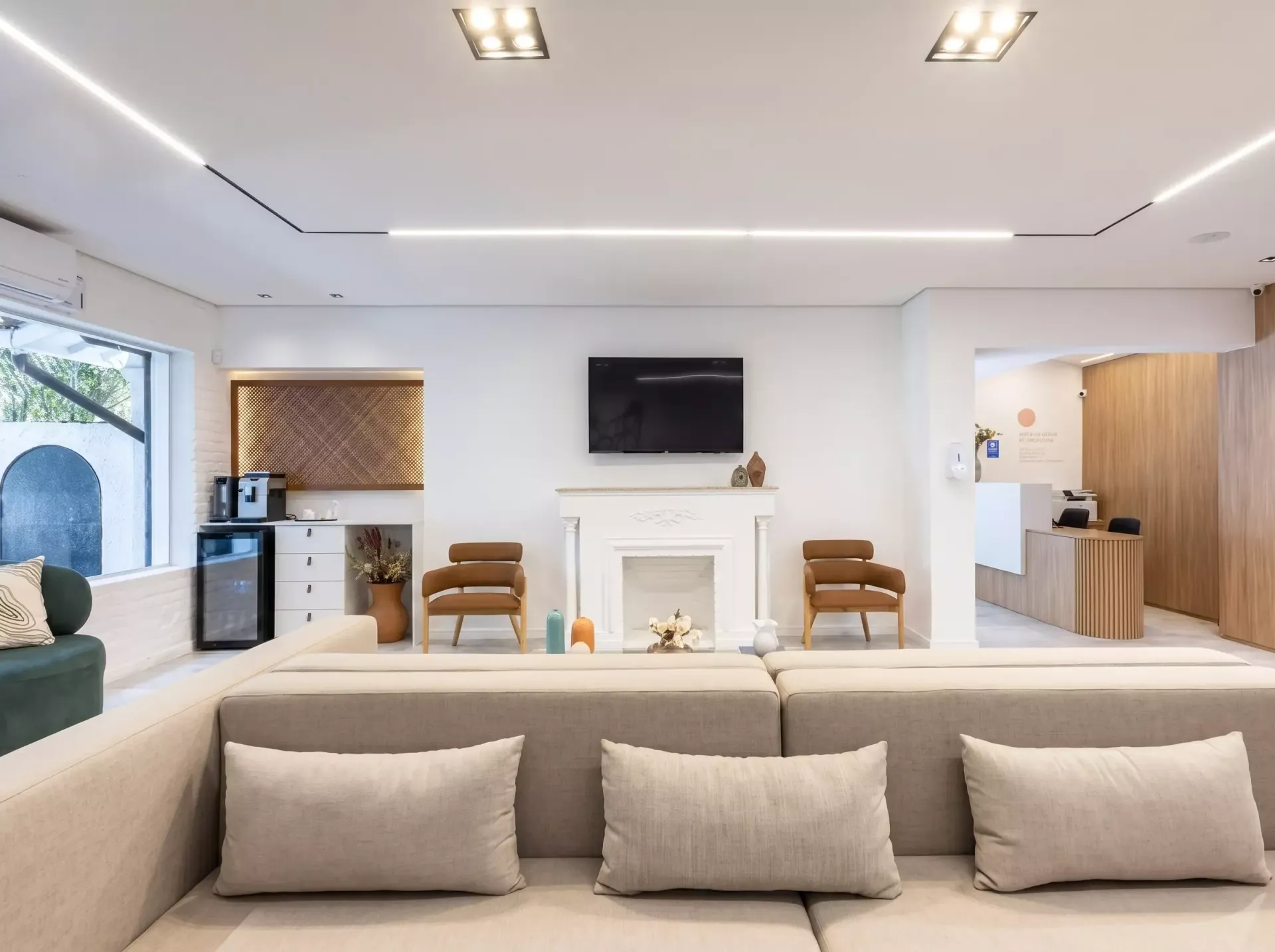 Sala de estar moderna e iluminada com lareira branca, TV de parede, sofá bege e detalhes em madeira.