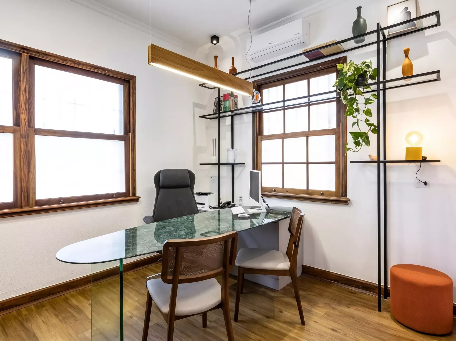 Escritório moderno em casa com mesa de vidro, cadeira, cadeira de escritório preta, prateleiras, plantas e janelas amplas.