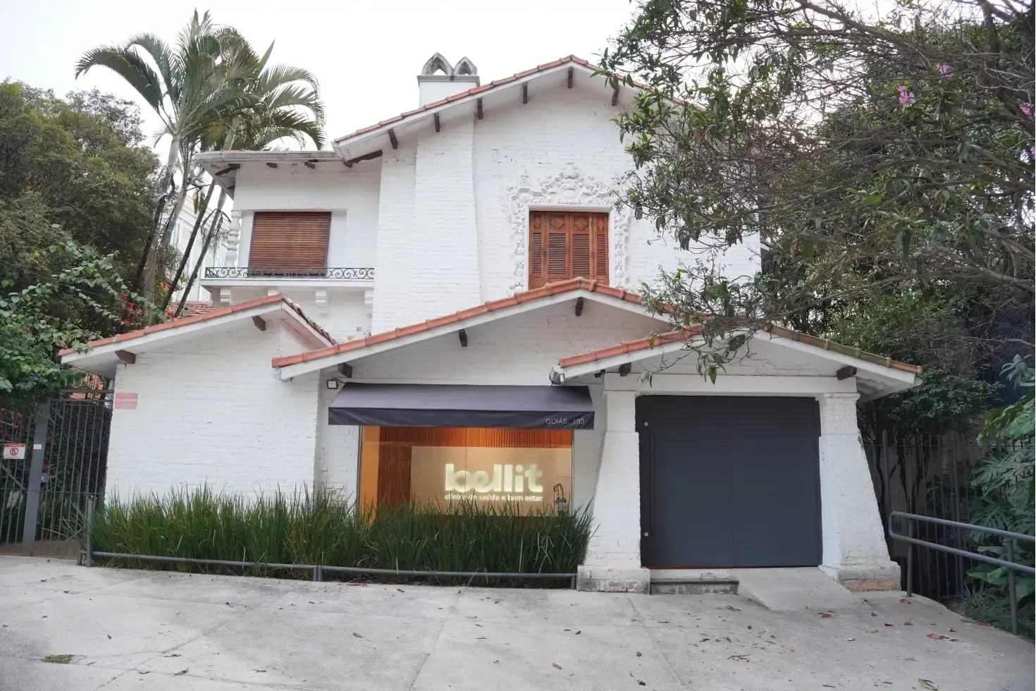Casa branca de dois andares com portão de garagem escuro, telhado de telhas e árvores na frente.
